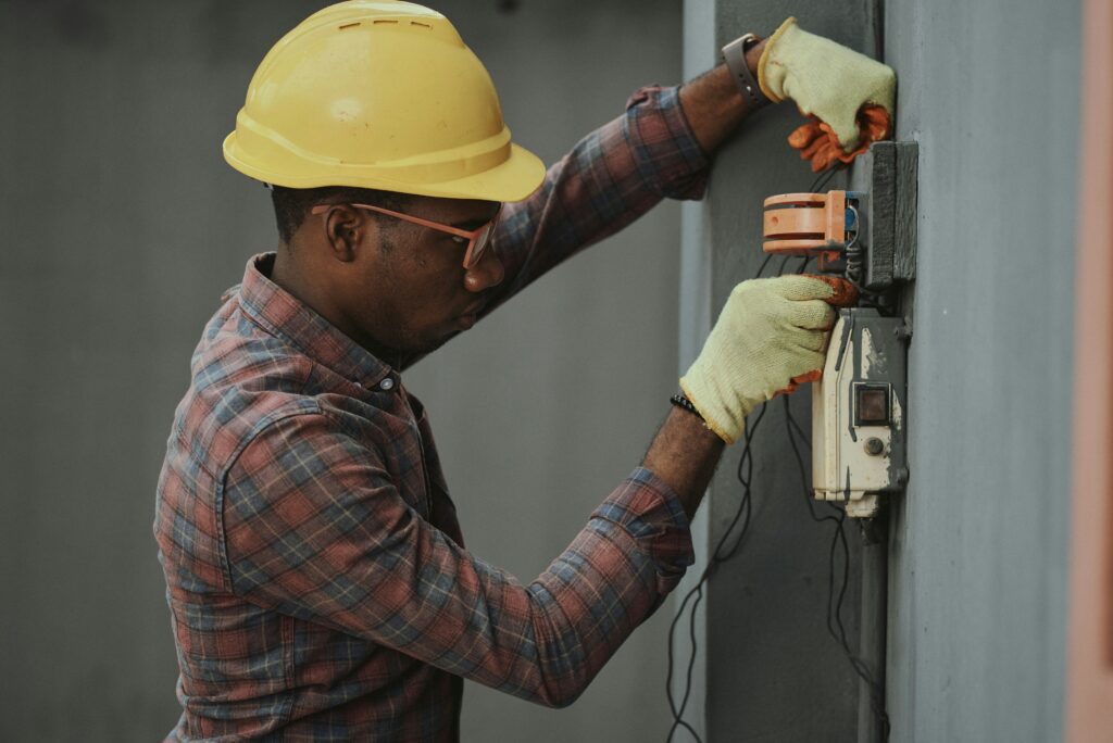 Electrician wearing a yellow hard hat and gloves working on electrical wiring outdoors.